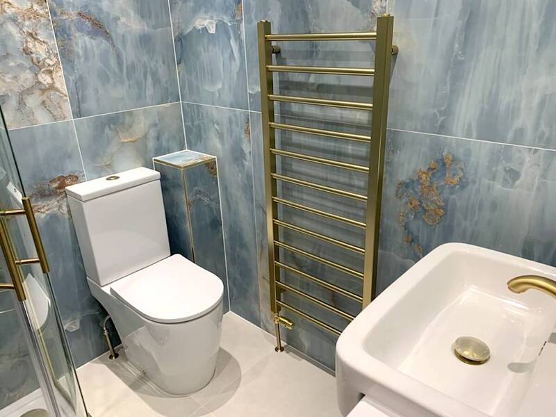 A close view of the same bathroom shows a white ceramic toilet with a concealed cistern, a small marble-effect shelf above it, and a gold-toned heated towel rail to the right. The wall is fully clad with marble-effect tiles, with varied blue and grey tones and a section of the glass shower enclosure is seen to the left