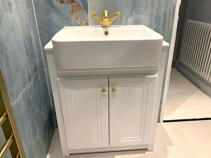 A modern, compact white bathroom vanity unit with a built-in basin, adorned with gold-toned fixtures, set against a wall of marble-effect ceramic tiles. The flooring appears to be light-coloured porcelain tiles, complementing the overall contemporary design. To the side, a small section of a gold-trimmed glass shower enclosure and a white radiator are visible