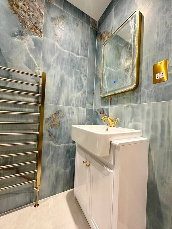 This image focuses on the vanity area, showcasing the white basin with gold tap, a matching gold-framed mirror above, and a gold switch plate to the side. The consistent marble-effect tile theme continues, and the heated towel rail can be seen to the left, enhancing the bathroom's luxurious feel