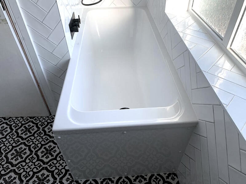 A pristine white bathtub boasts a sleek design, perfectly complemented by a herringbone tile arrangement and ornate floor tiles, brightening the space with natural light from the window