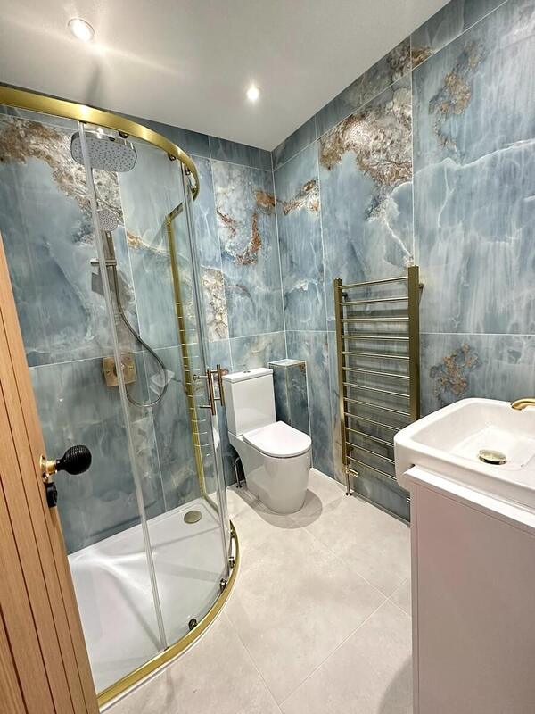 A wider perspective of the bathroom reveals the full shower enclosure with gold trim, the white toilet, and the vanity unit. The consistent marble-effect tiling covers all walls and the floor is laid with light-coloured tiles. Two recessed ceiling lights provide illumination